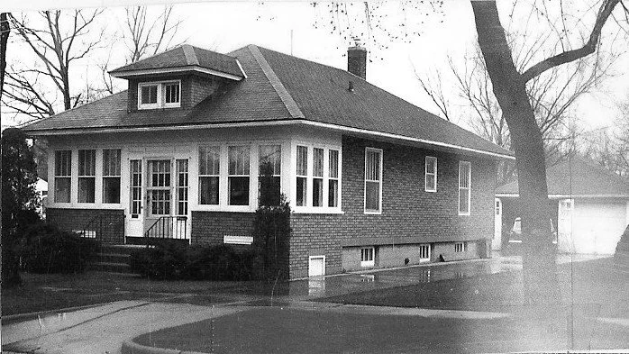Black-and-white photo of a one-story brick bungalow with an enclosed front porch, leafless trees, and a driveway leading to a detached garage.