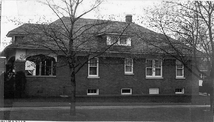 Black-and-white photo of a one-story brick bungalow with a covered front porch, dormer windows, and leafless trees along the sidewalk.
