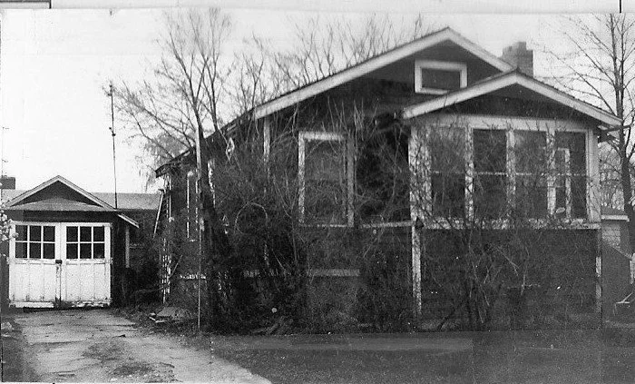 Black-and-white photo of an older one-story bungalow with overgrown bare shrubs and trees, and a detached garage to the left.
