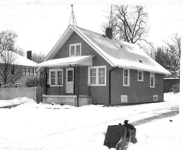 Black-and-white photo of a small single-story bungalow with a covered front porch and snow-covered yard, a tied trash bag sits in the foreground.