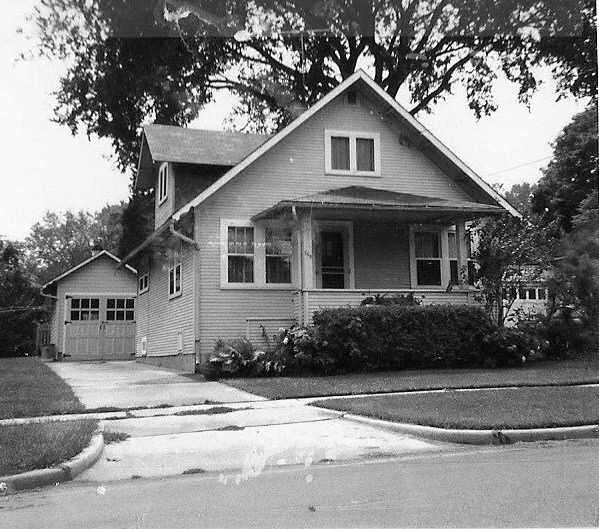 Black-and-white photo of a two-story Craftsman-style house with a covered front porch, detached garage to the left, and a large tree behind it.