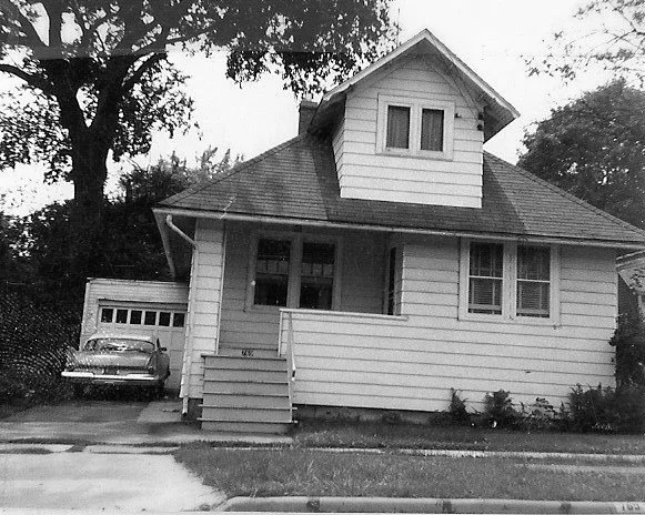 Black-and-white photo of a small two-story house with a covered front porch, steps leading up, a detached garage to the left with a vintage car parked beside it, and a large tree in the background.