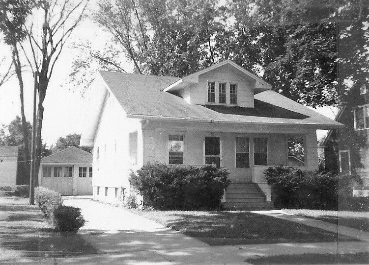 Black-and-white photo of a single-story bungalow with a front porch, shrubs, driveway and detached garage.
