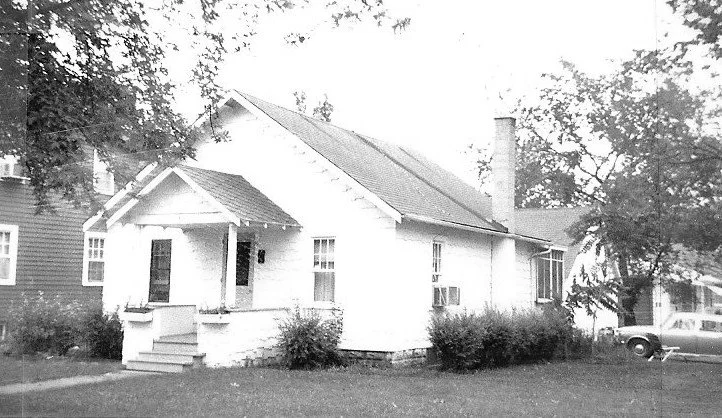Black-and-white photo of a small single-story house with a covered front porch, chimney, shrubs in the yard, and a car parked to the right.