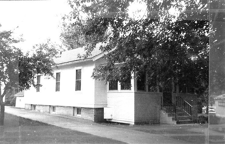 Black-and-white photo of a single-story wooden house with an enclosed front porch, front steps, and trees shading the yard.