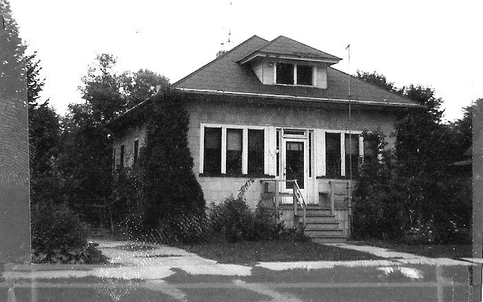 Black-and-white photo of a single-story bungalow with an enclosed front porch, central doorway reached by a short flight of steps, and tall shrubs and trees on either side.