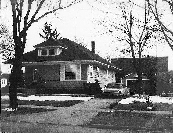 Black-and-white photo of a single-story bungalow with a car in the driveway, bare trees and patches of snow on the lawn.