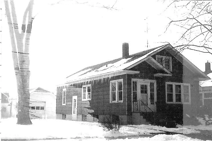 Black-and-white photo of a single-story house in winter with icicles hanging from the roof, bare trees, a detached garage, and snow covering the yard.