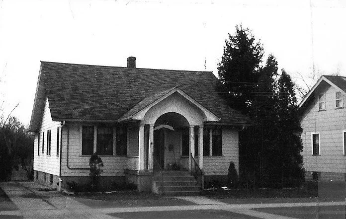 Black-and-white photo of a single-story bungalow with a covered front porch and steps, flanked by a large evergreen tree and a neighboring house.