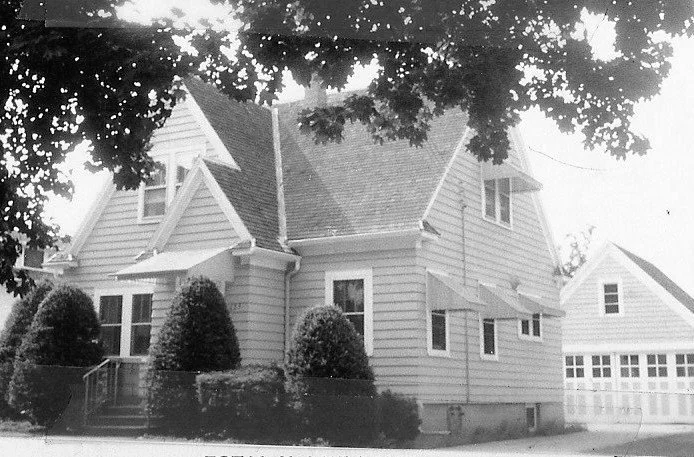 Black-and-white photo of a two-story wood-sided house with trimmed shrubs in front, a detached garage to the right, and tree branches framing the top.