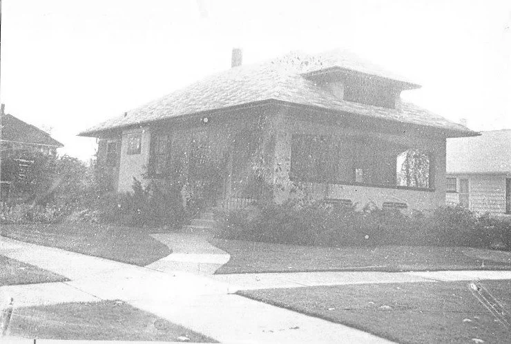 Black-and-white photo of a single-story bungalow with a wide covered porch, front steps, lawn, and intersecting sidewalks.