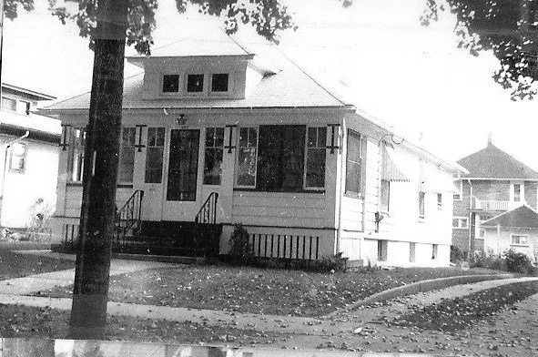 Black-and-white photo of a single-story bungalow with a screened front porch, two short stairways, and a tree in the foreground.
