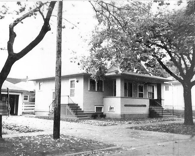 Black-and-white photo of a single-story brick bungalow with a covered front porch, two staircases, a detached garage to the left, and large trees and a utility pole in the front yard.