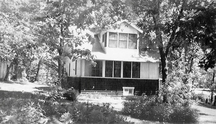 Black-and-white photo of a two-story house with an enclosed front porch, shaded by large trees and surrounded by a grassy, fern-filled yard.