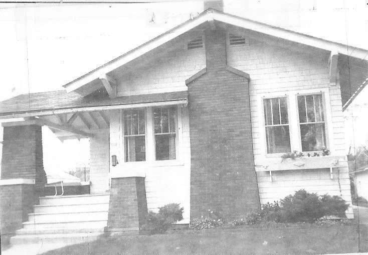 Front view of a single-story Craftsman bungalow with a centered brick chimney, covered porch, front steps, and double-hung windows.