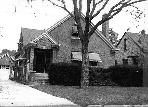 Black-and-white photo of a brick bungalow with awnings, a large tree and shrubs in the front yard, and a driveway leading to a detached garage.