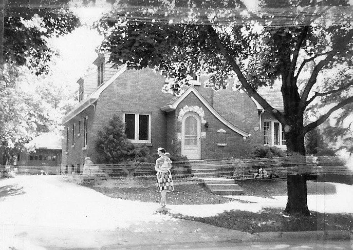 Black-and-white photo of a brick cottage with an arched front door, large shade tree, and a woman in a patterned dress standing on the lawn.