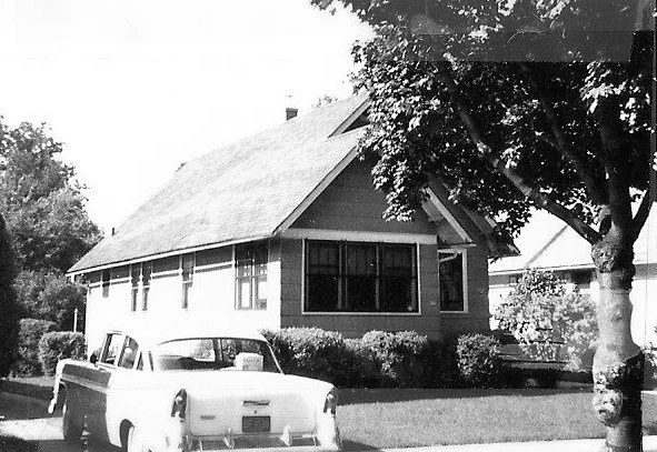 Black-and-white photo of a small bungalow with shrubs, a large tree in front, and a 1950s-era car parked on the street.