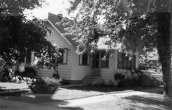 Black-and-white photo of a small single-story house with a front porch and steps, shaded by large trees and surrounded by a lawn and shrubs.