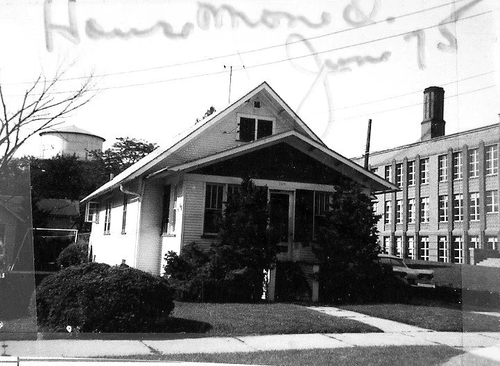Black-and-white photo of a small bungalow with front shrubs and a sidewalk; a multi-story brick factory with a smokestack and a water tower are visible in the background.