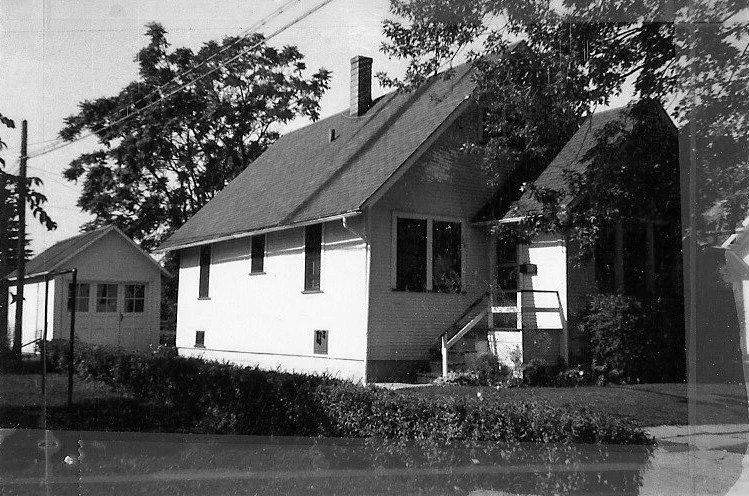 Black-and-white photo of a small gable-roof house with front steps, a detached garage to the left, and trees around.