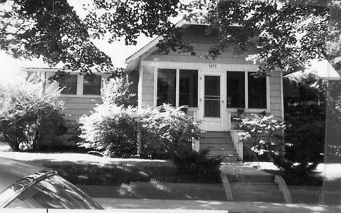 Black-and-white photo of a small bungalow with screened front porch, house number "1073" above the door, shrubs and large shade trees, and a car hood visible in the lower-left foreground.