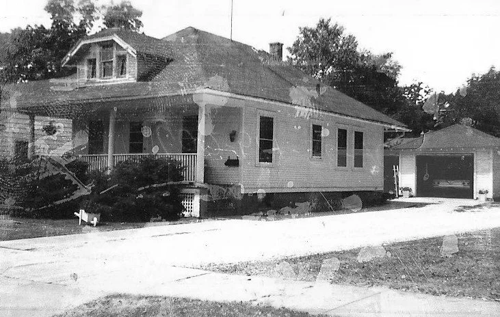 Black-and-white photo of a single-story bungalow with a covered front porch and columns, driveway to a detached garage, and trees in the background.