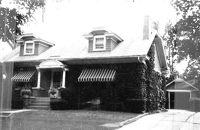 Black-and-white photo of a one-and-a-half-story house with two dormer windows, striped window awnings, vine-covered facade, front steps with planters, and a driveway leading to a detached garage.