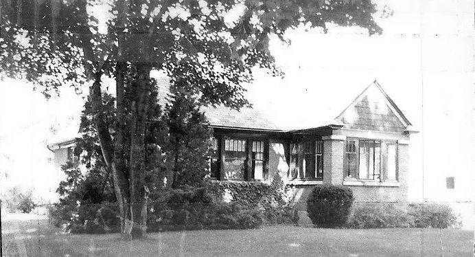 Black-and-white photo of a single-story bungalow-style house partially shaded by large trees, with shrubs and a lawn in the front.