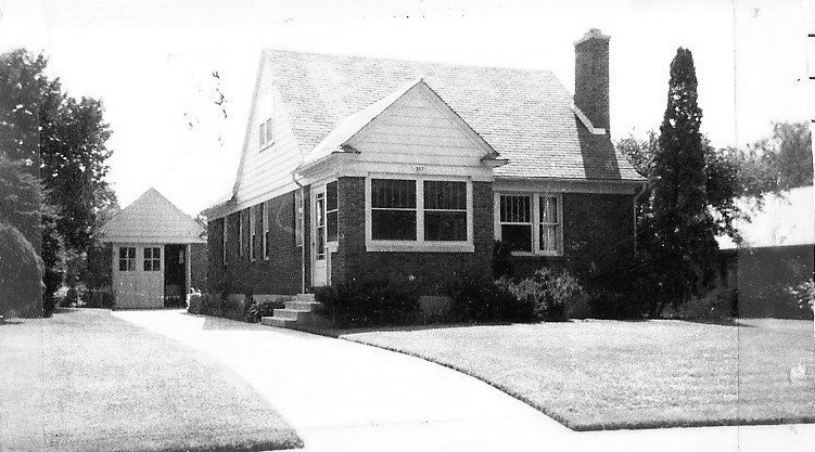 Black-and-white photo of a single-story brick house with a front lawn, curved driveway leading to a detached garage, and a tall chimney.