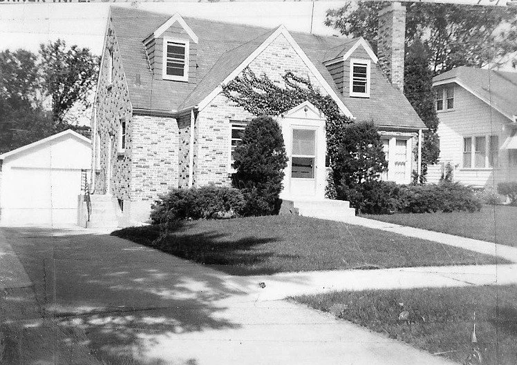 Black-and-white photo of a brick Cape Cod–style house with two dormer windows, a centered front door flanked by shrubs, a sidewalk and driveway leading to a detached garage at left.