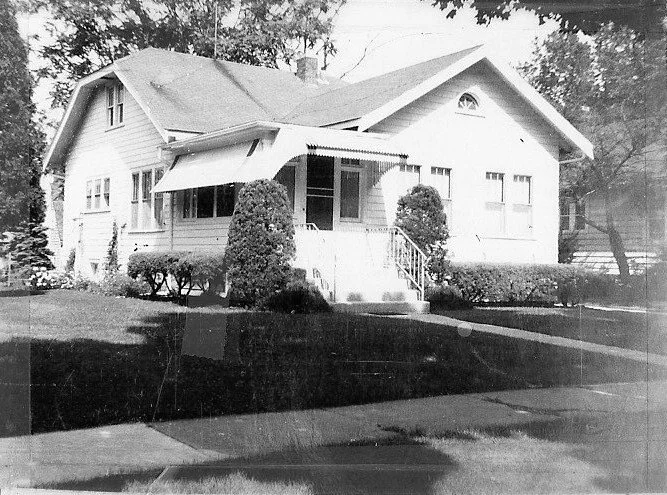 Black-and-white photo of a single-story suburban house with a covered front porch, awning, shrubs, and a front lawn.