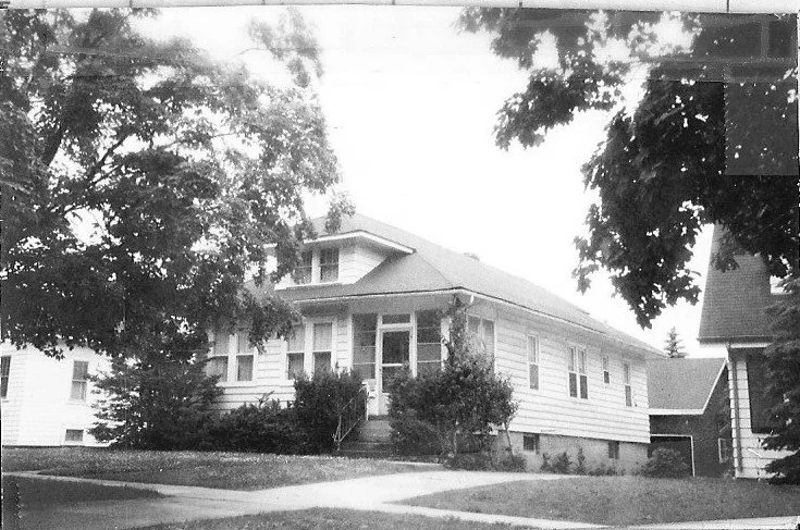 Black-and-white photo of a single-story bungalow with an enclosed front porch, trees and a sidewalk.