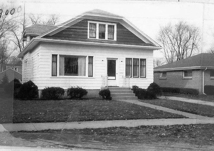 Black-and-white photo of a two-story bungalow-style house with front steps, shrubs, and a sidewalk.