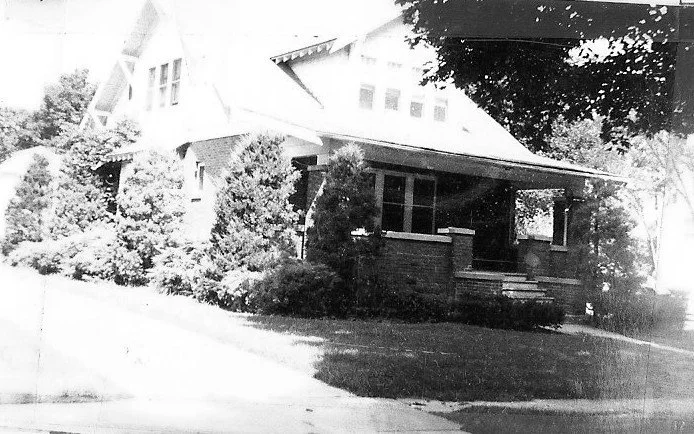 Black-and-white photo of a Craftsman-style brick bungalow with a covered front porch, large shrubs and trees, and a sloping lawn leading to a sidewalk.