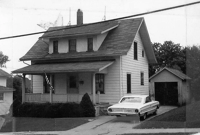 Black-and-white photo of a two-story clapboard house with a covered front porch and American flag, an older sedan parked in the driveway beside a detached garage.