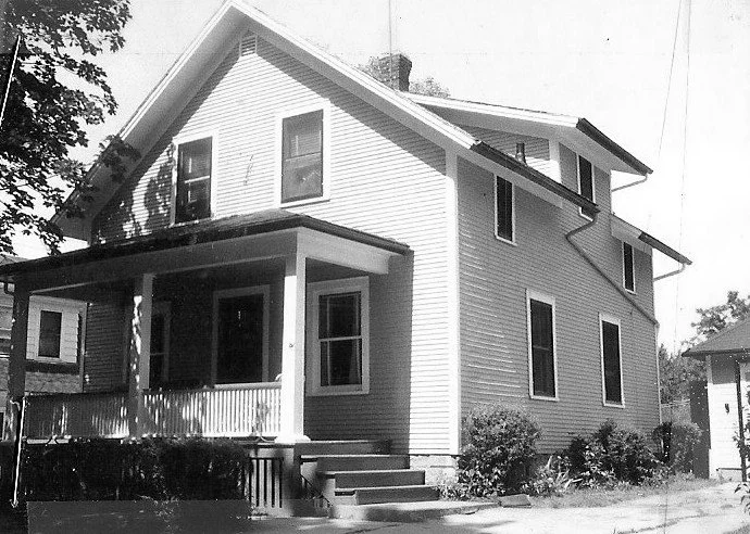 Black-and-white photo of a two-story gabled house with a front porch, steps, and shrubs.