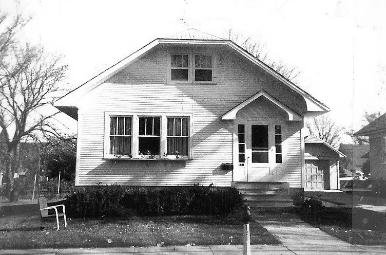 Black-and-white photo of a small bungalow with a front porch, three front windows, detached garage to the right, and a front lawn.
