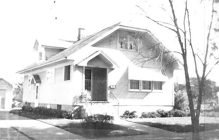 A black-and-white photo of a two-story Craftsman-style house with a covered front porch, sidewalk and small yard with trees.