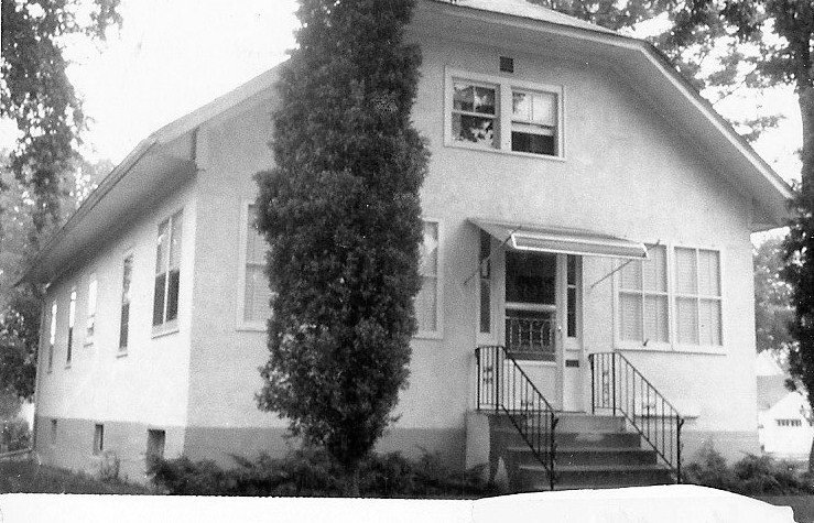 Black-and-white photo of a small stucco bungalow with front steps, metal awning, and a tall narrow evergreen tree beside the entrance.