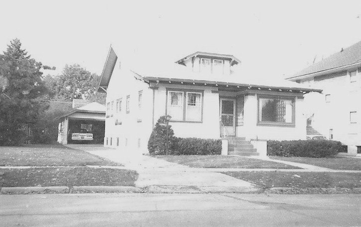 Front view of a single-story bungalow with a small front porch, shrubs and lawn, and a driveway leading to a carport with a parked car.