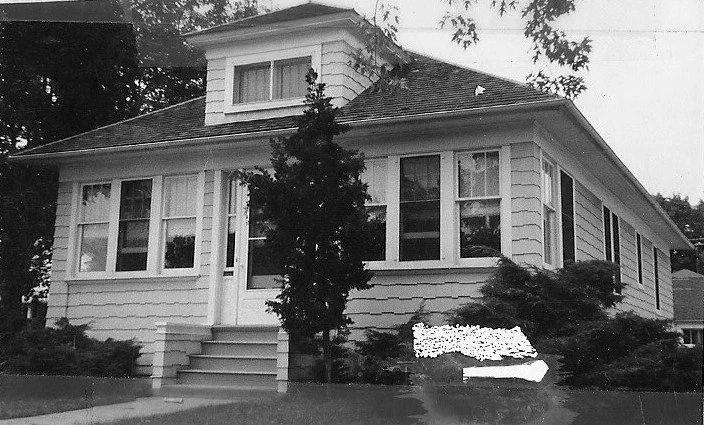 Black-and-white photo of a one-story bungalow with front steps, a central dormer window, shrubs, and a small tree.