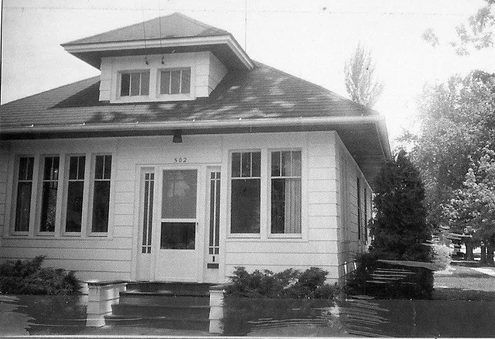 Black-and-white photo of a single-story bungalow with a central front door, tall windows, and a small dormer on the roof.