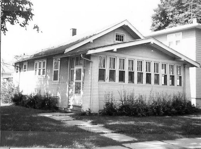 Black-and-white photo of a single-story bungalow with an enclosed front porch of many windows, a small lawn and a sidewalk leading to the front door.