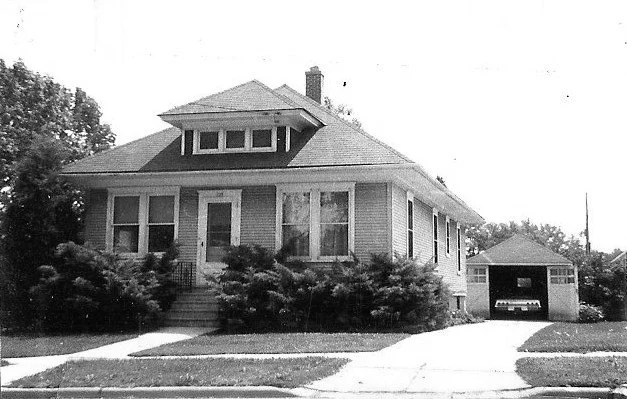 Black-and-white photo of a one-story bungalow with a front porch, shrubs along the facade, and a detached garage with a car.