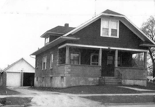 A black and white photo of a house with a front porch and a detached garage.
