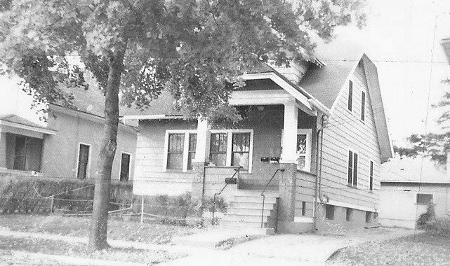 Black-and-white photo of a single-family bungalow with a front porch and steps, large tree in the yard, and a driveway leading to a detached garage.
