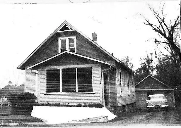 Black-and-white photo of a wood-sided bungalow with an enclosed front porch, detached garage to the right and a vintage car inside.