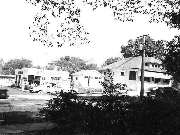 Black-and-white photo of a small Standard gas station with parked cars next to residential buildings, framed by overhanging tree branches.