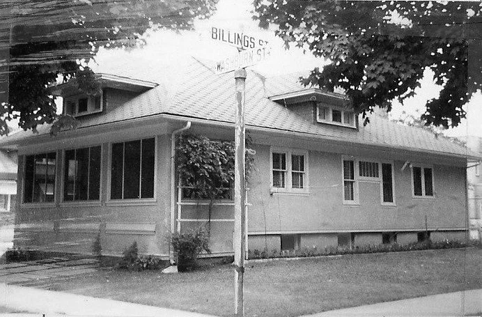 Black-and-white photo of a single-story bungalow on a tree-lined corner lot with a porch and a street sign reading "BILLINGS" and "WASHINGTON ST."